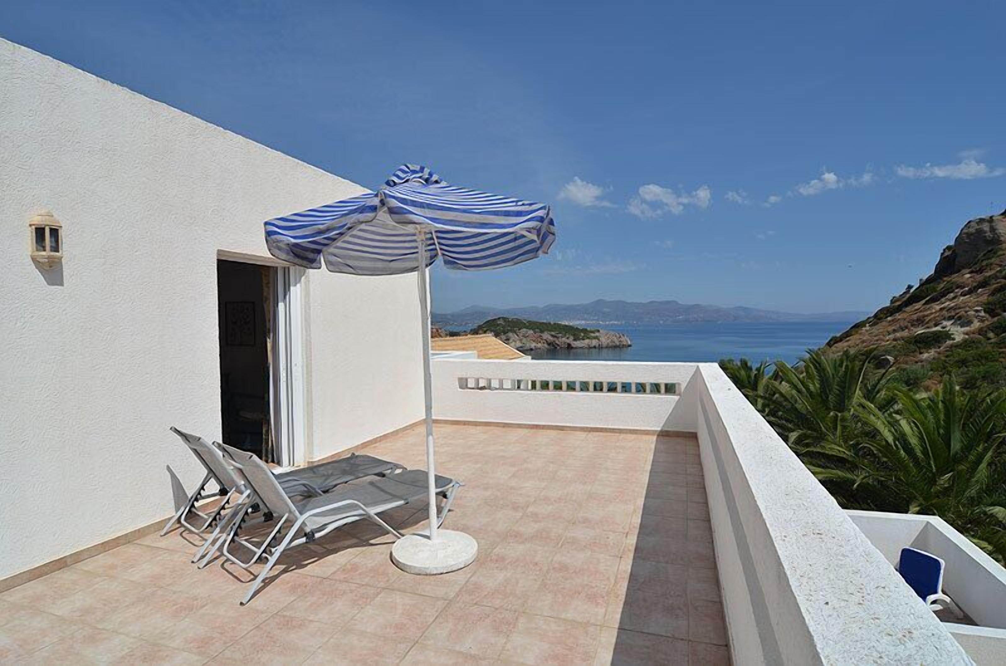A terrace with a white wall, a blue-striped umbrella, two lounge chairs, and a view of the sea.