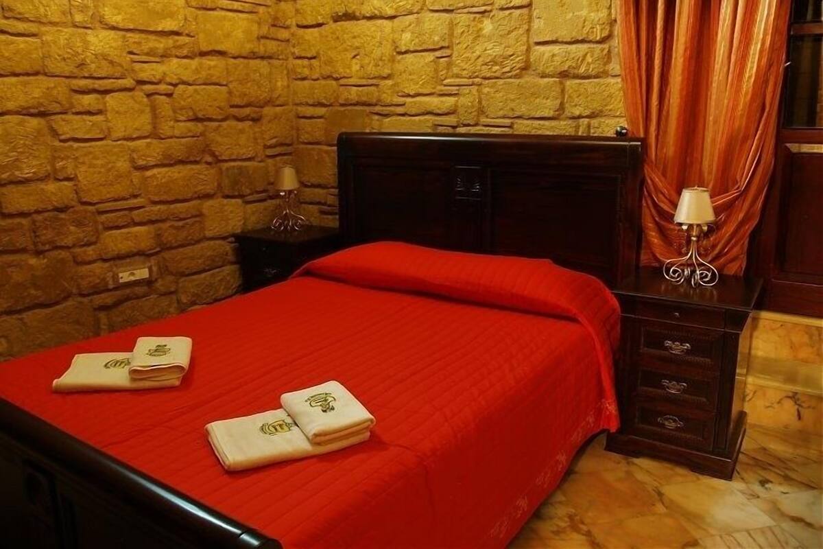 Une chambre avec un mur en pierre, une tête de lit en bois, une couverture rouge et deux serviettes pliées sur le lit.
