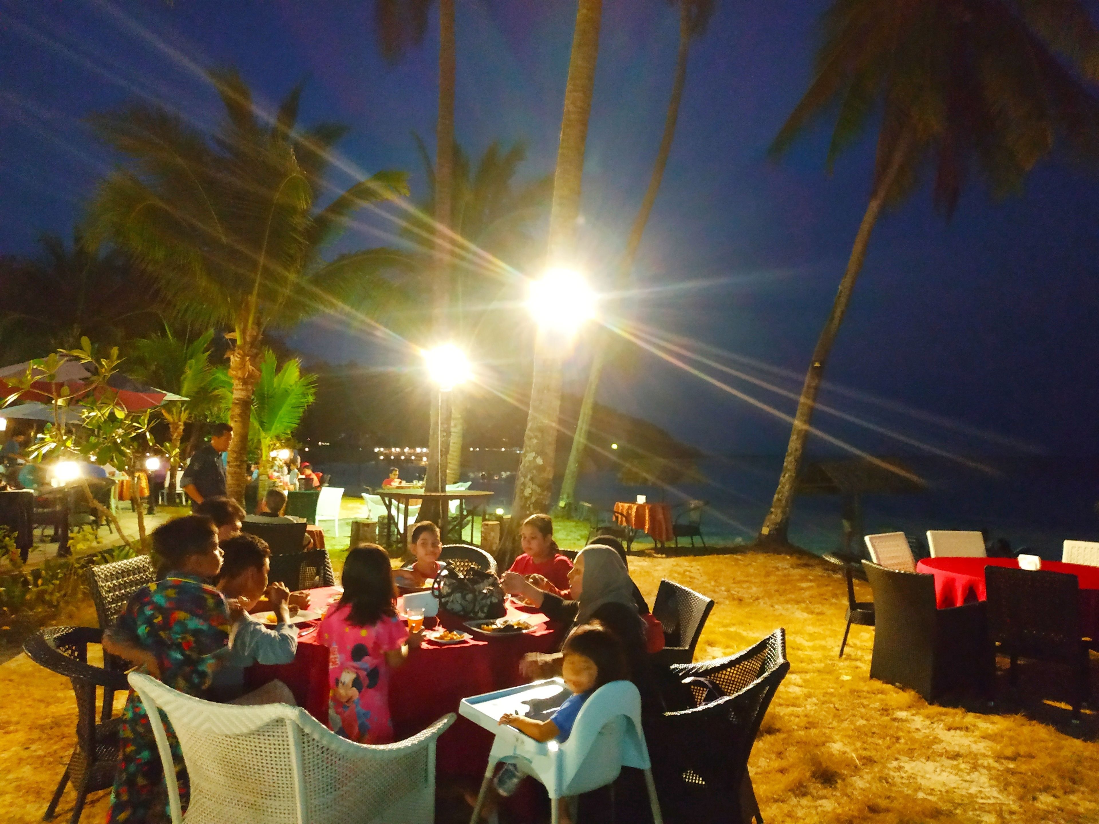 An outdoor dining area with tables and chairs, palm trees, and a beach in the background.