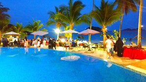 Área de piscina com palmeiras, guarda-sóis e pessoas.