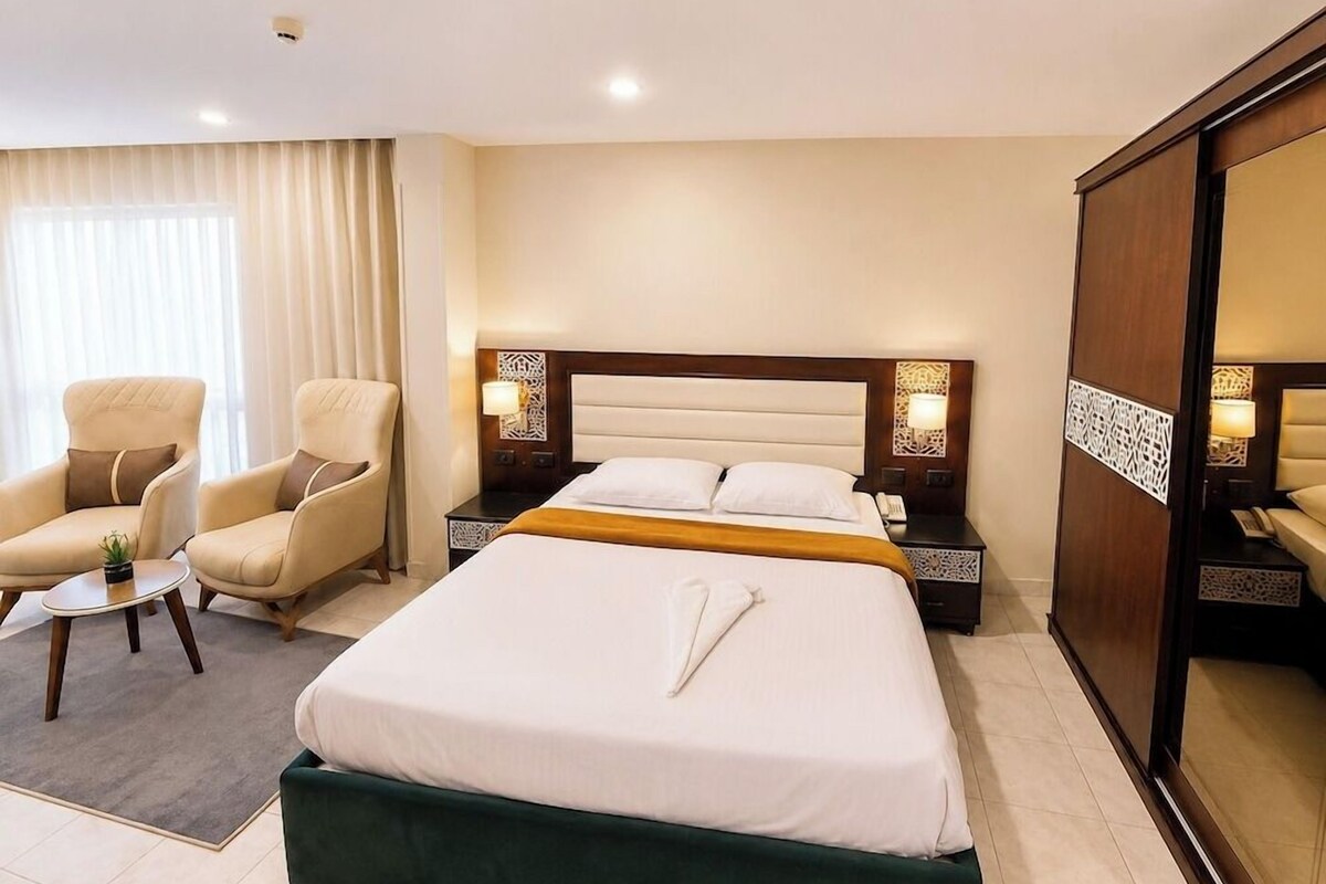 A hotel room with a large bed, two armchairs, a small table, and a wardrobe.