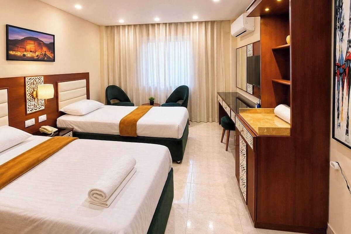 A hotel room with two beds, a desk, a chair, a TV, and a painting on the wall.