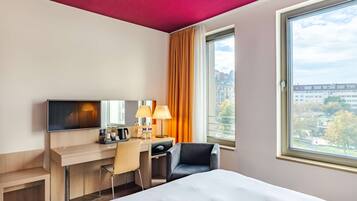 A hotel room with a bed, desk, chair, TV, and a window with a city view.