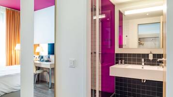 Habitación de hotel con cama, escritorio, silla y baño con espejo grande, lavamanos y cabina de ducha con azulejos morados.