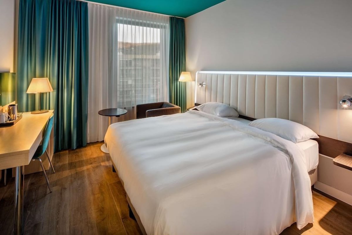 A hotel room with a large bed, a desk with a chair, a small table, and a window with curtains.