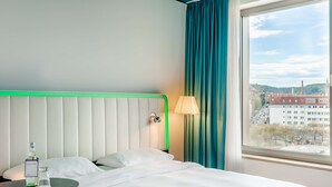 A hotel room with a bed, a bedside table with a bottle and glass, a lamp, and a window with a view of buildings and trees.
