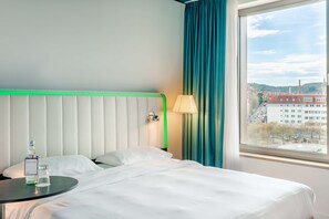 A hotel room with a bed, a bedside table with a bottle and glass, a lamp, and a window with a view of buildings and trees.