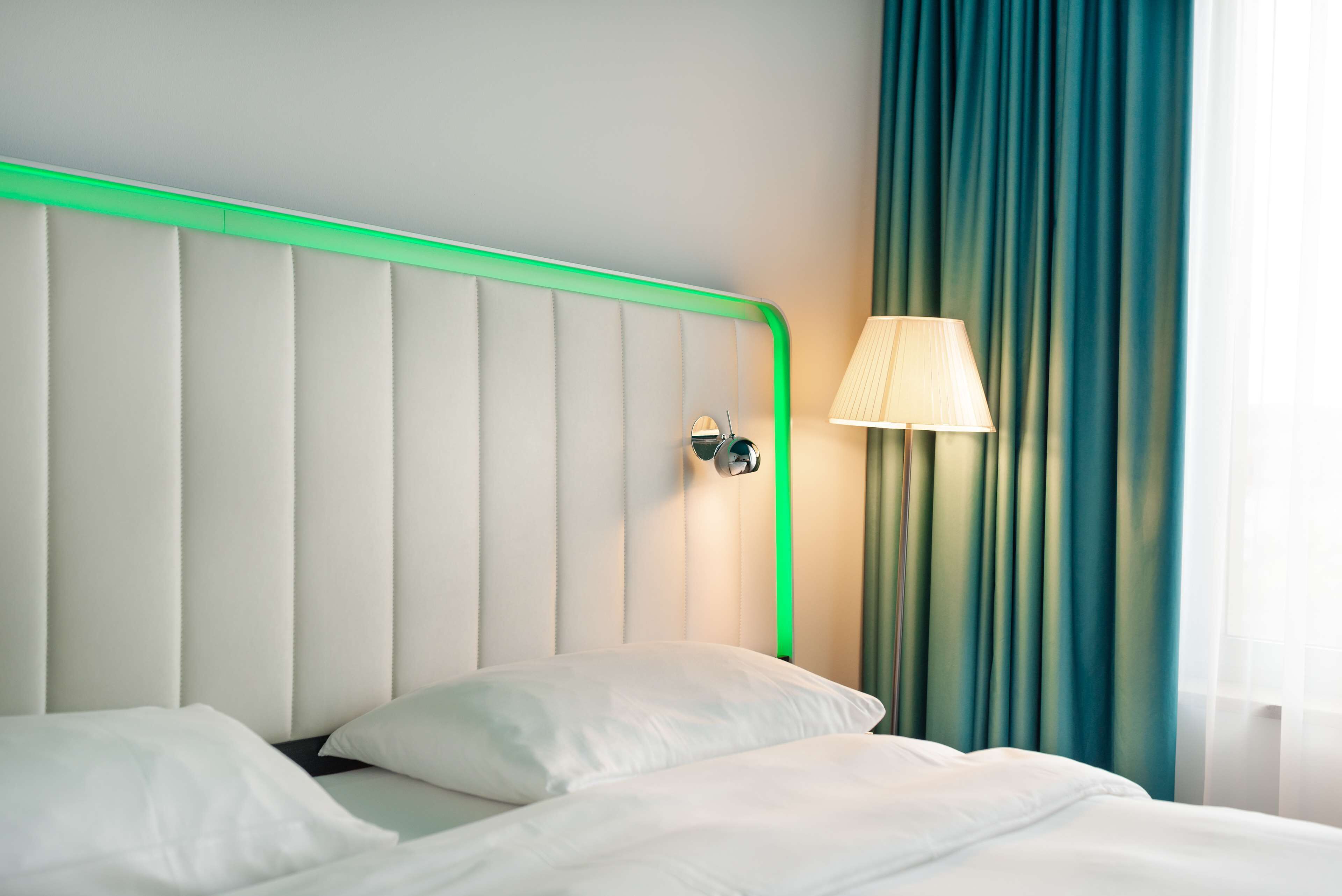 Una habitación de hotel con una cama, cabecero con iluminación verde, lámpara y cortinas azul verdosas.