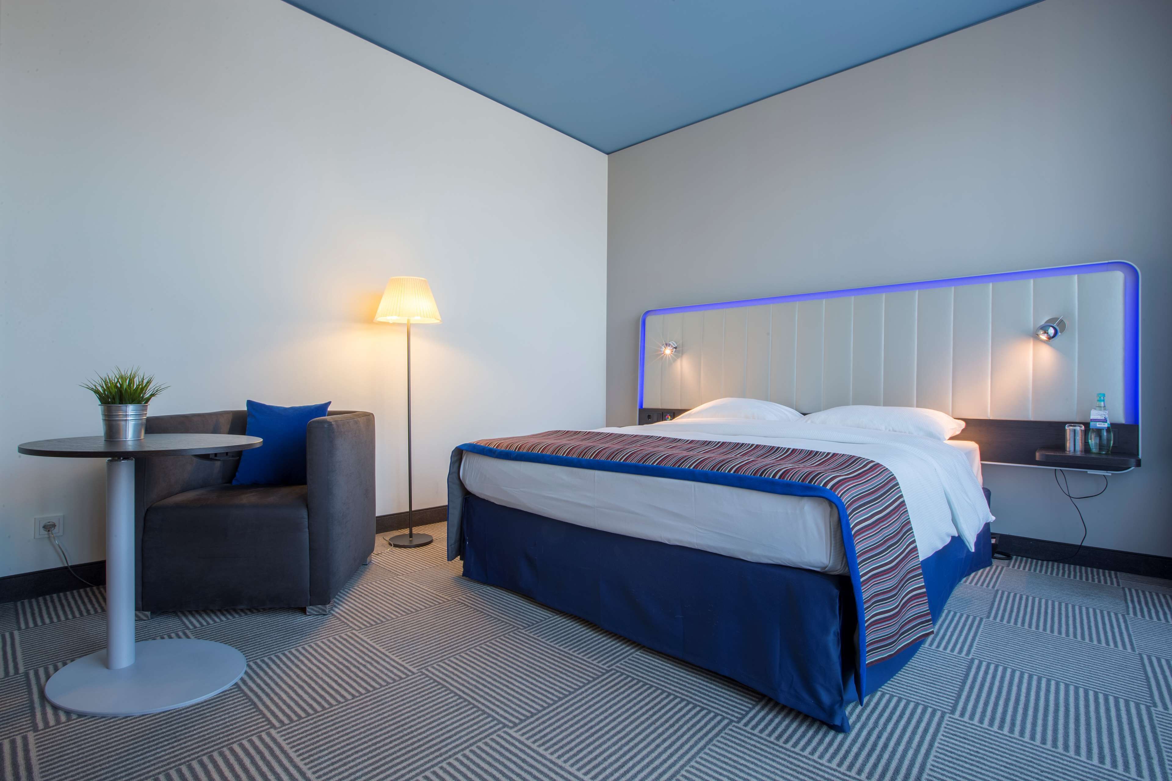 Una habitación de hotel con una cama, un escritorio, una silla, una lámpara y una mesita con una planta.