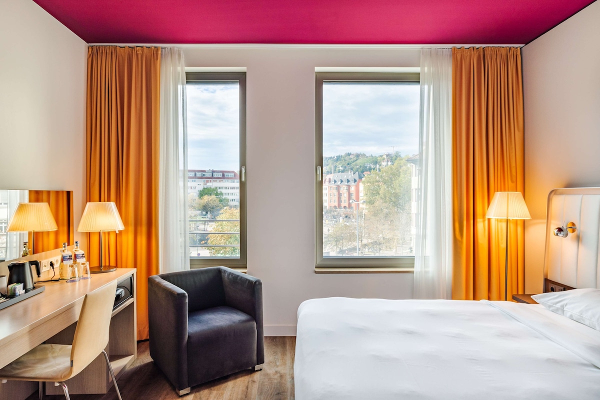 A hotel room with a bed, desk, chair, two windows, and a view of the city.