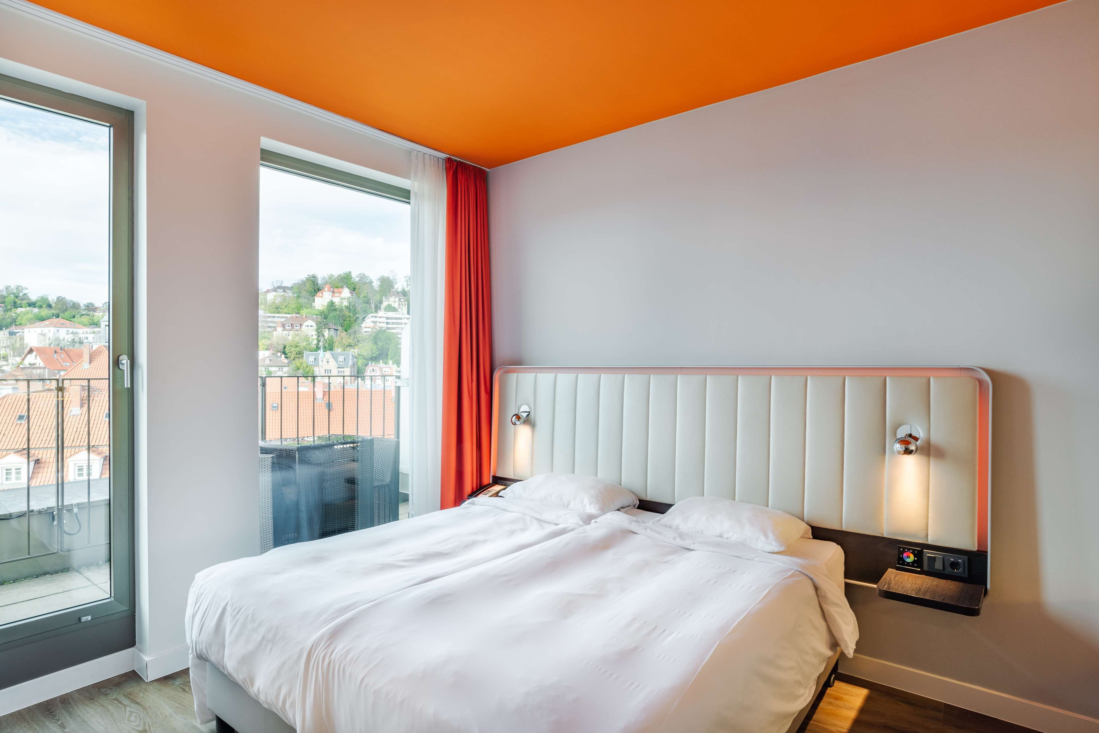 A hotel room with a large window, a bed with white linens, a red curtain, and a white headboard with wall-mounted lights.