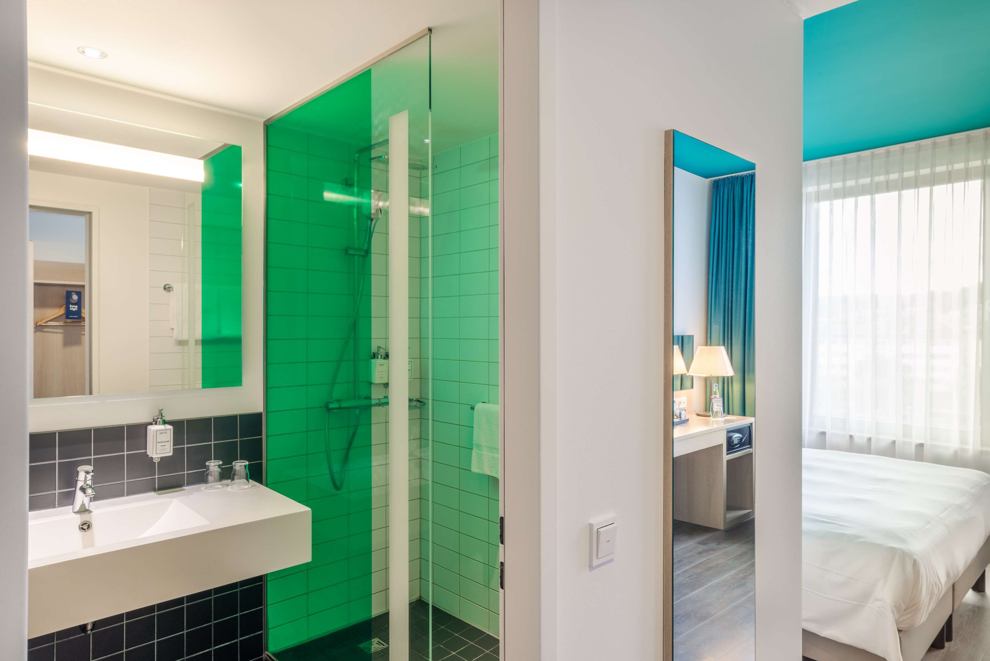 A modern hotel room with a bathroom featuring a green-tiled shower, a white sink with a mirror, and a bed with a blue accent wall.