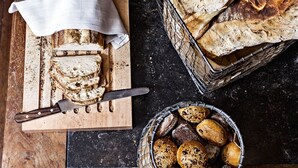 A wooden cutting board with sliced bread and a knife, a basket of assorted breads, and a cloth napkin on a dark countertop.
