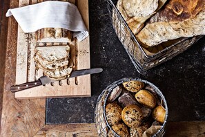 A wooden cutting board with sliced bread and a knife, a basket of assorted breads, and a cloth napkin on a dark countertop.