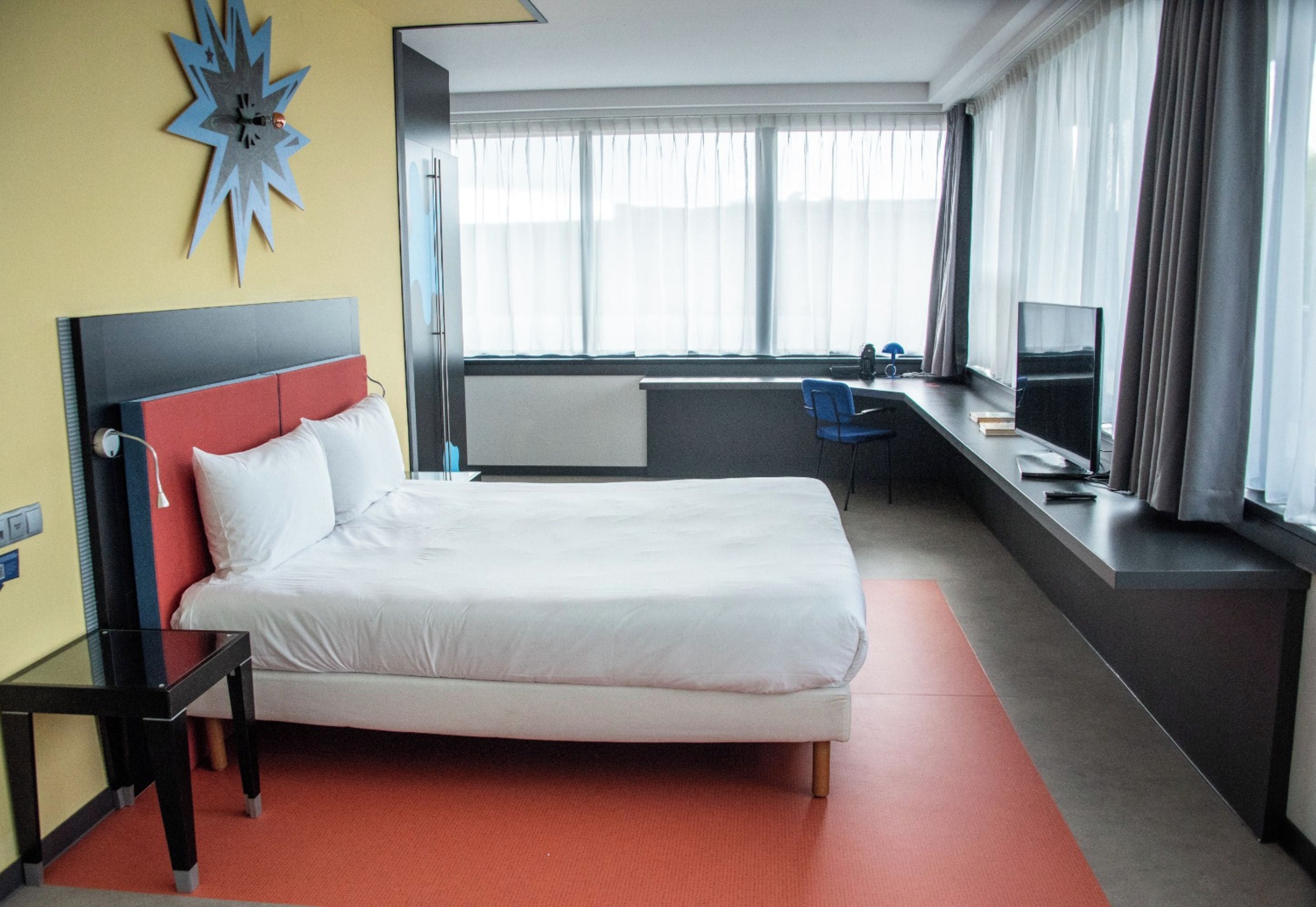 Habitación de hotel con cama, escritorio, silla, televisor y ventana con cortinas.