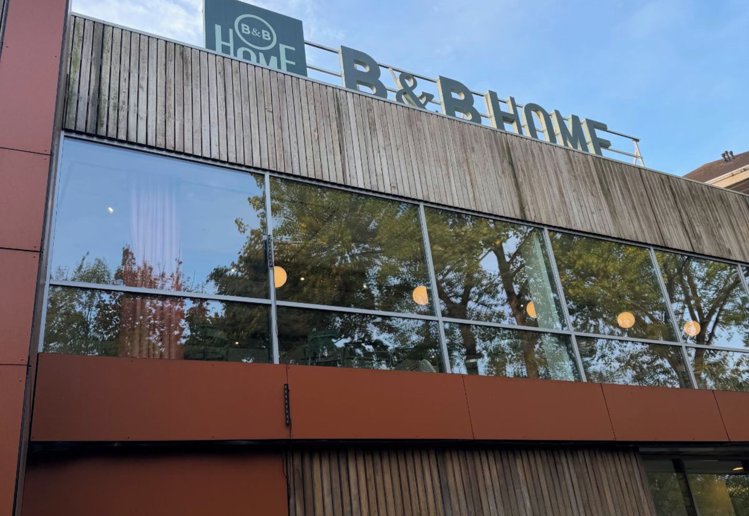 Un bâtiment doté d’une grande enseigne indiquant « B&B HOME » et présentant un design architectural moderne avec de grandes fenêtres qui reflètent les arbres.