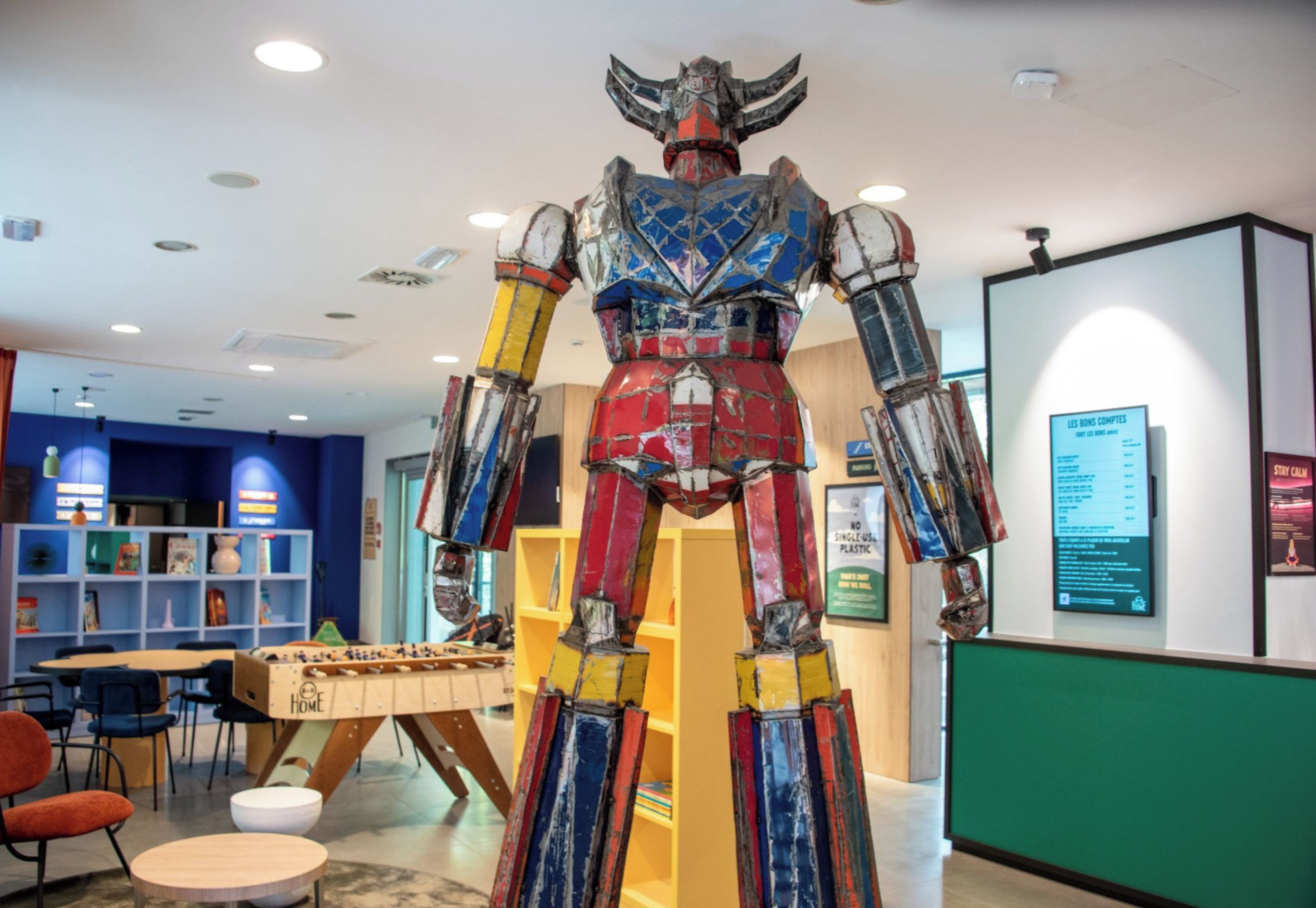 A colorful sculpture of a humanoid figure in a public space with bookshelves and tables in the background.