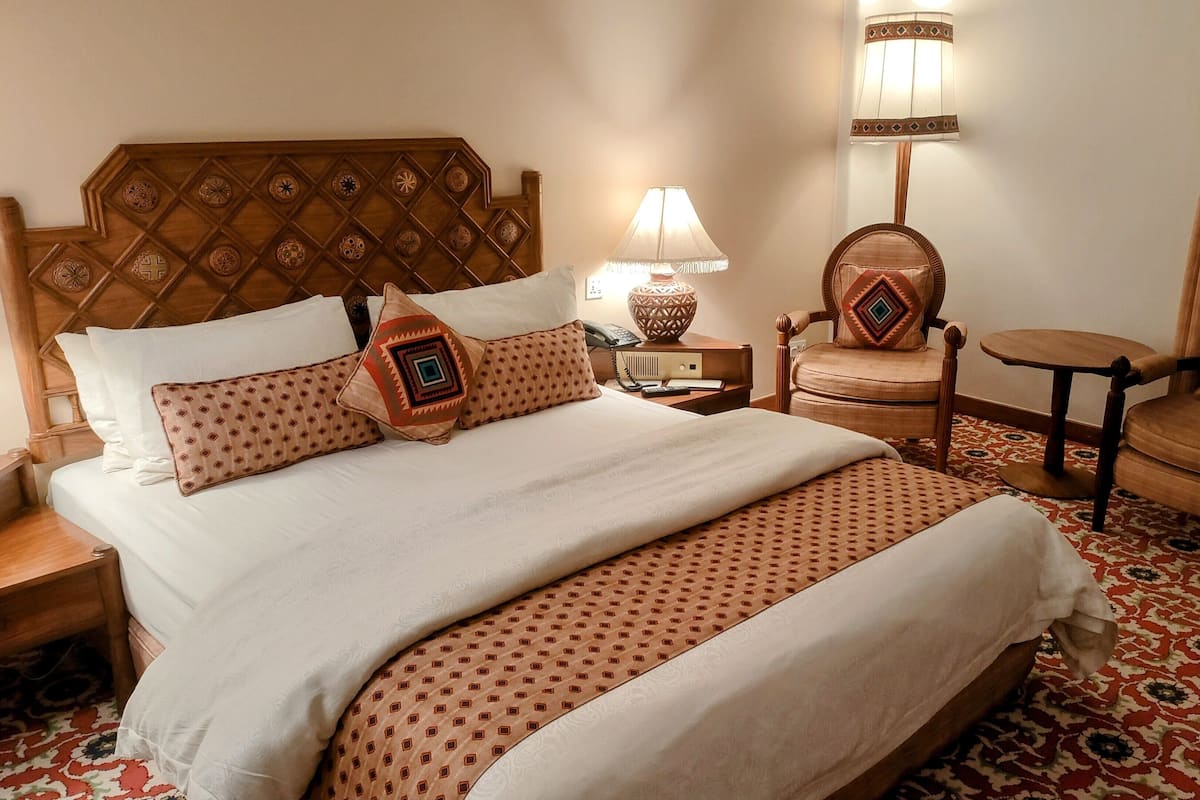 A hotel room with a large bed, two chairs, a small table, a lamp, and a patterned carpet.