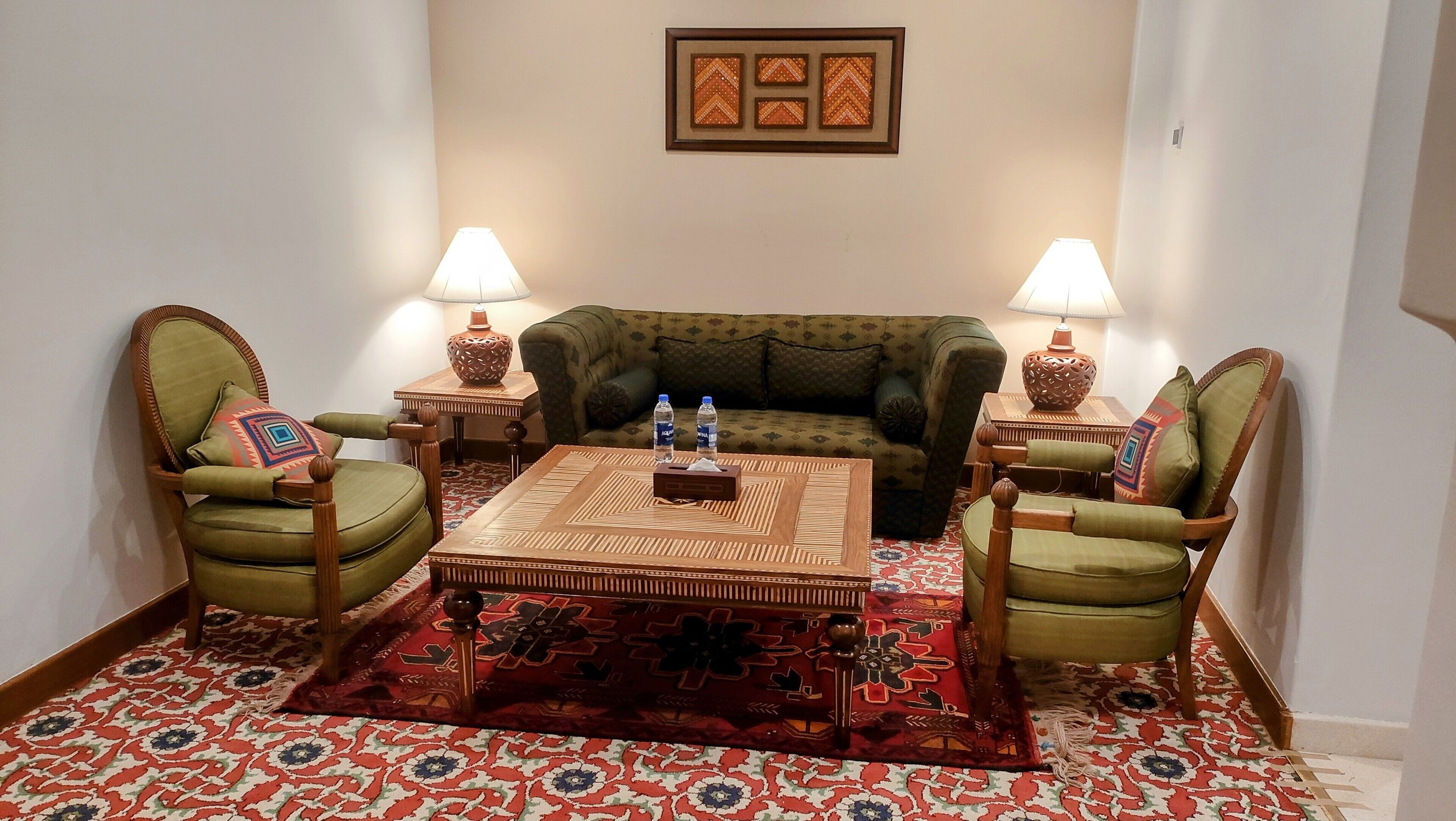 Ein Wohnzimmer mit einem grünen Sofa, zwei Sesseln, einem Couchtisch und zwei Lampen. Auf dem Sofa stehen zwei Wasserflaschen.