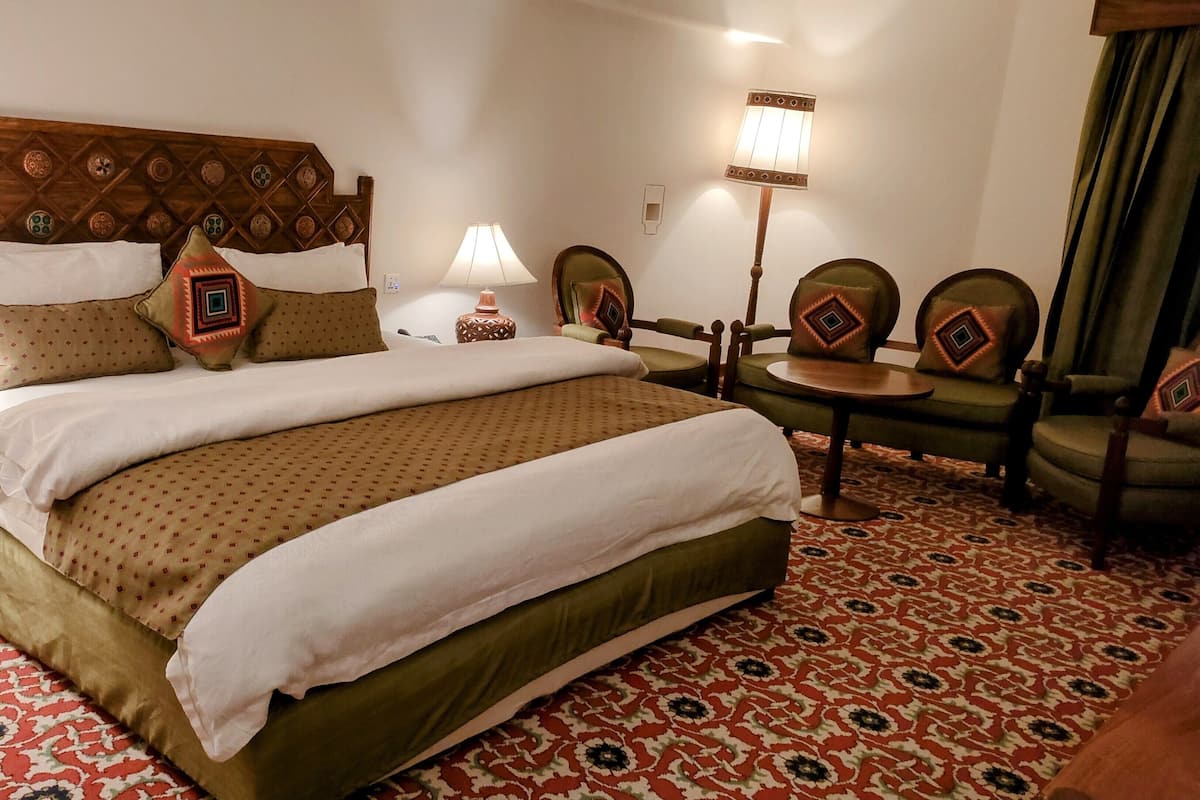 A hotel room with a large bed, two bedside tables, a lamp, a chair, and a patterned carpet.