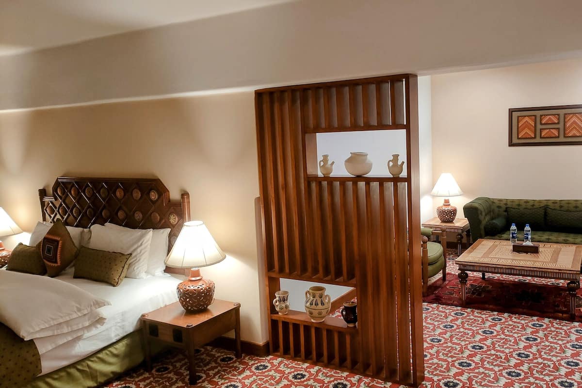 A hotel room with a bed, a wooden partition, a green sofa, a coffee table, and a patterned carpet.