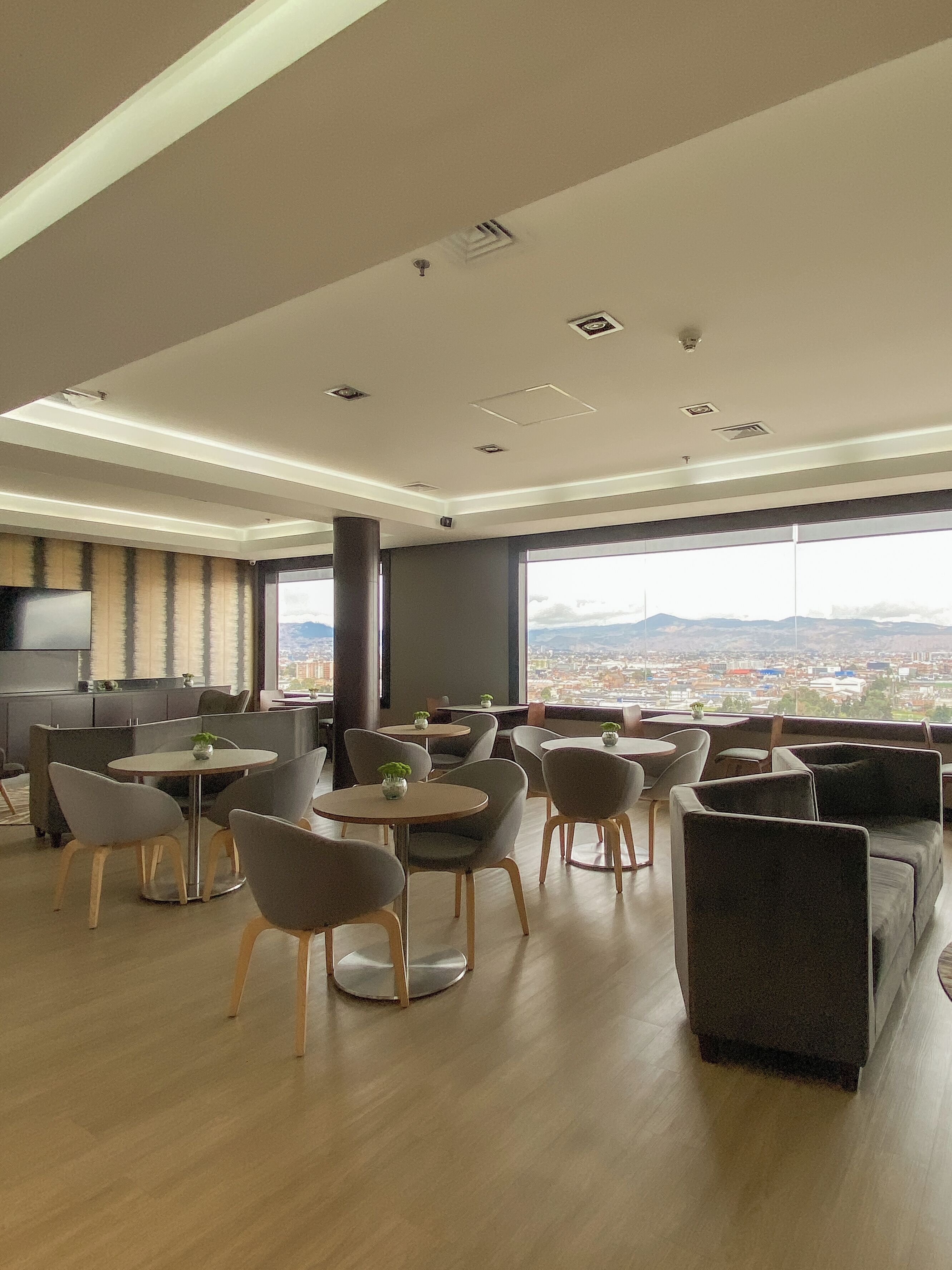 Un comedor moderno con mesas y sillas redondas, ventanas amplias que ofrecen vista a la ciudad y un televisor de pantalla plana montado en la pared.