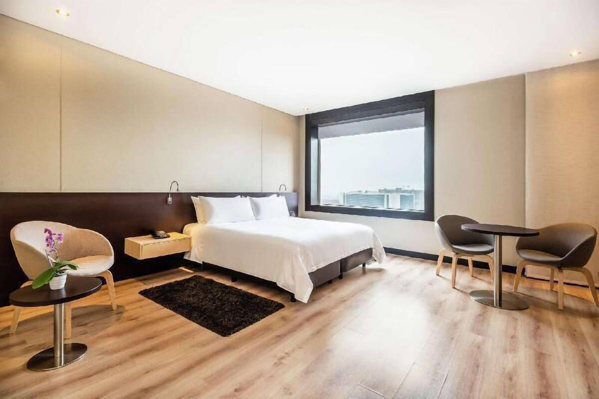 A modern hotel room with a bed, a desk, a chair, and a large window.