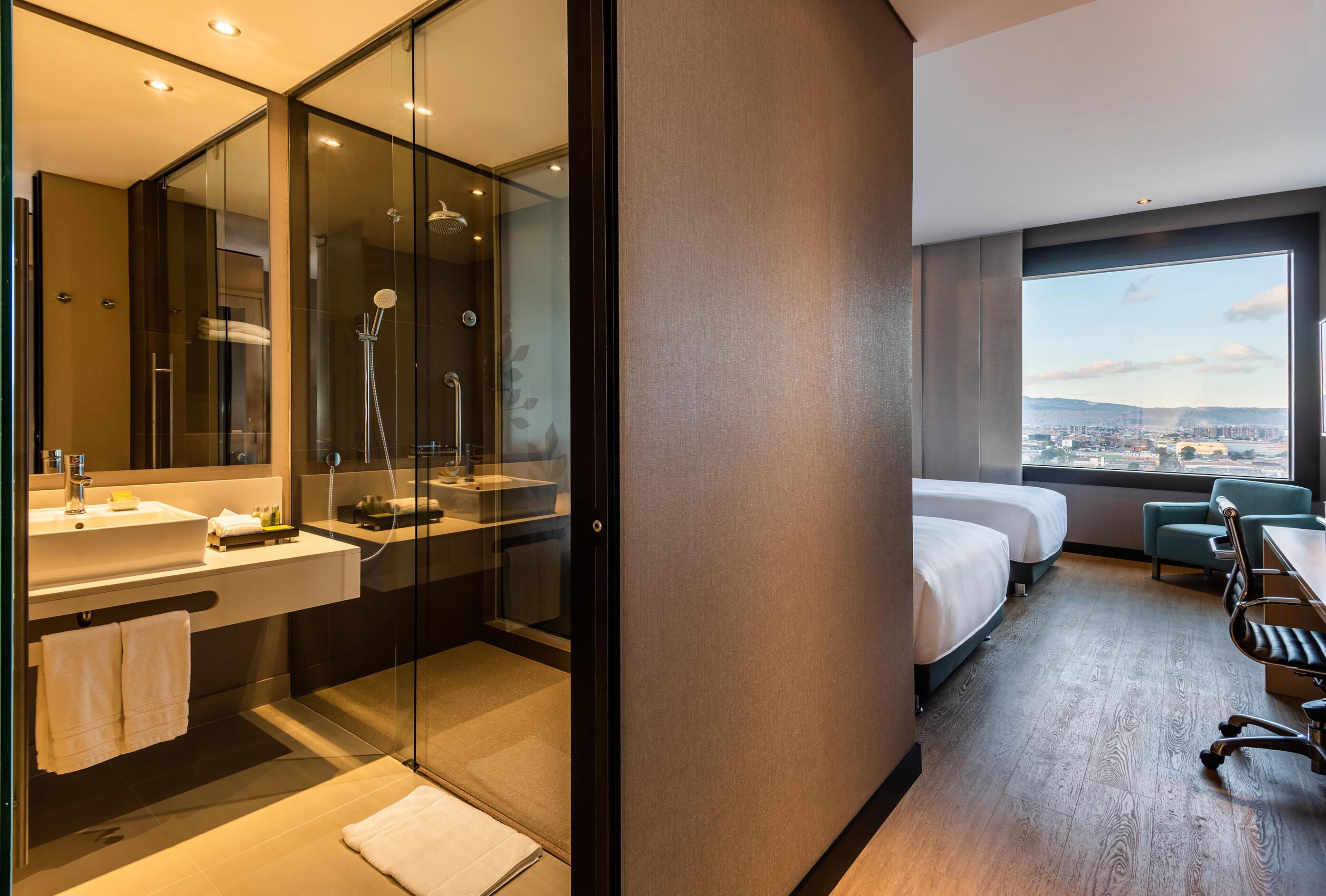 A modern hotel room with a glass-enclosed shower, a large bed, a desk, and a view of the city.