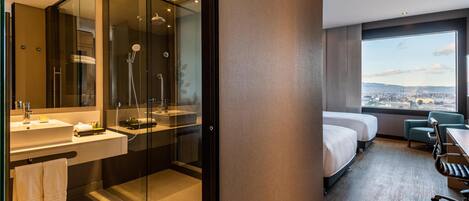 A modern hotel room with a glass-enclosed shower, a large bed, a desk, and a view of the city.