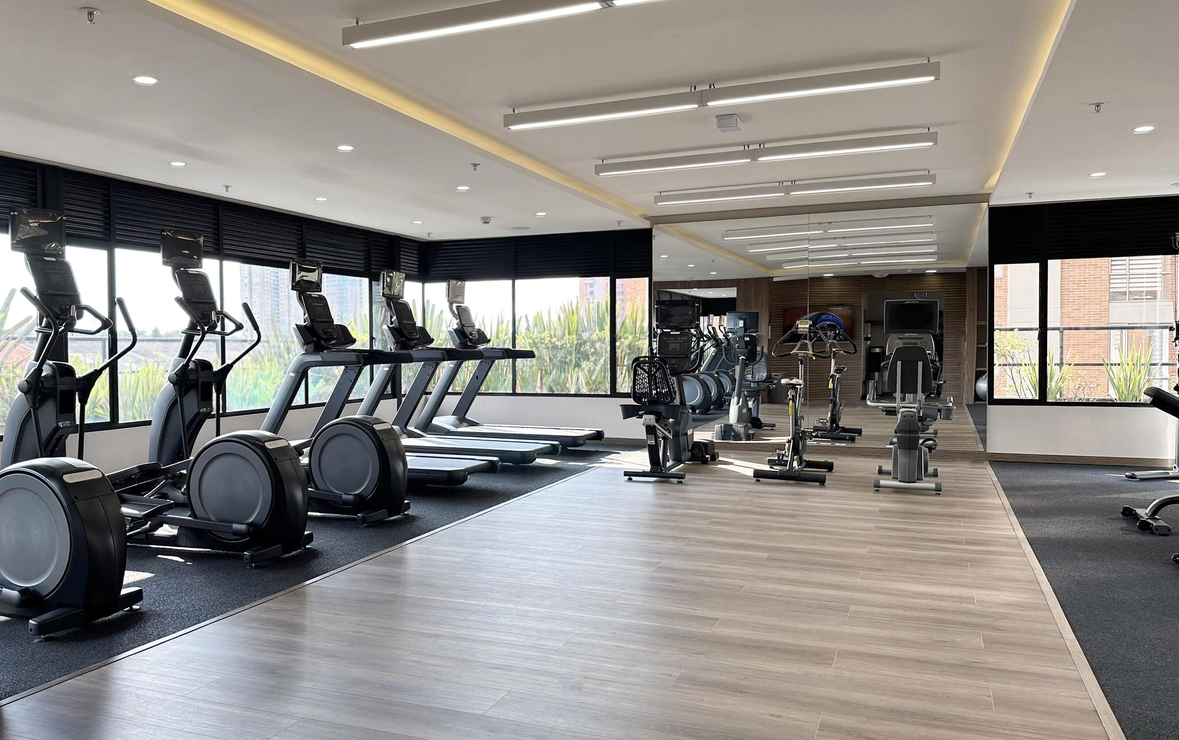 Une salle de sport moderne équipée de divers appareils de cardio, d’un mur avec des miroirs et de grandes fenêtres donnant sur un paysage verdoyant.