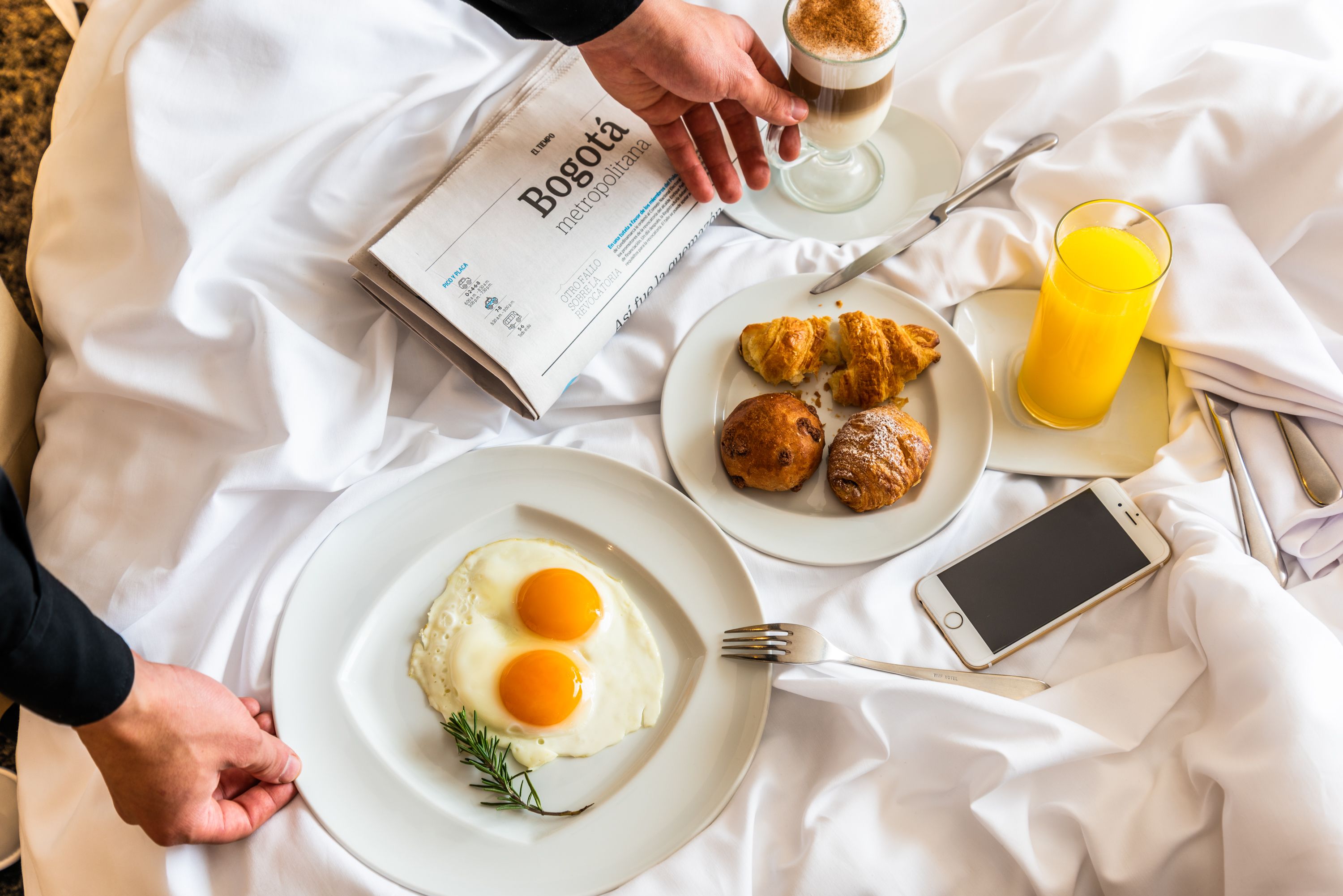 Desayuno con periódico, huevos, cruasanes y un vaso de jugo de naranja sobre un mantel blanco.