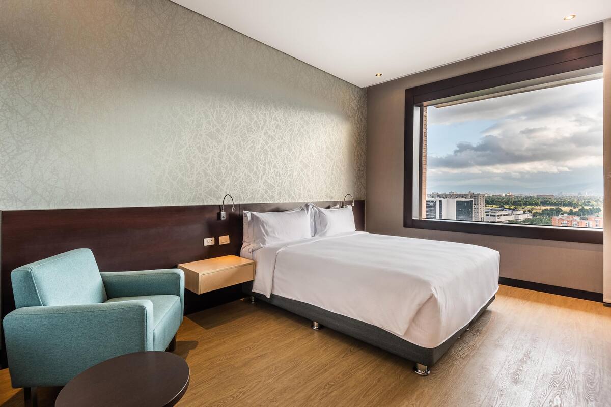 A hotel room with a bed, a chair, a nightstand, and a large window with a city view.
