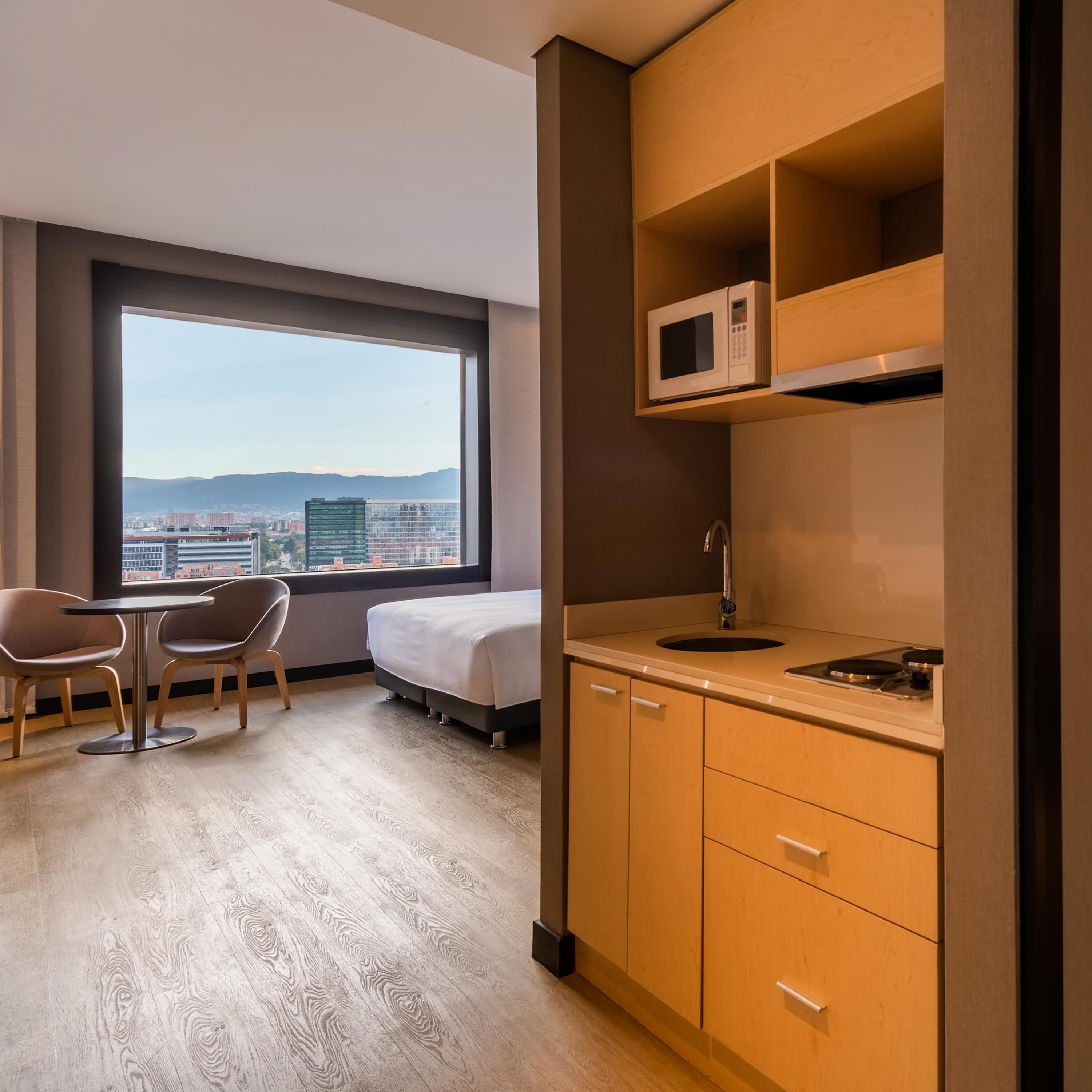 Una habitación de hotel con cama, una kitchenette pequeña, un comedor con sillas y un ventanal con vistas a la ciudad.