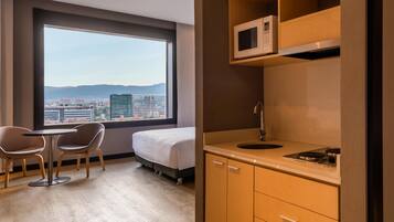 Una habitación de hotel con cama, una kitchenette pequeña, un comedor con sillas y un ventanal con vistas a la ciudad.