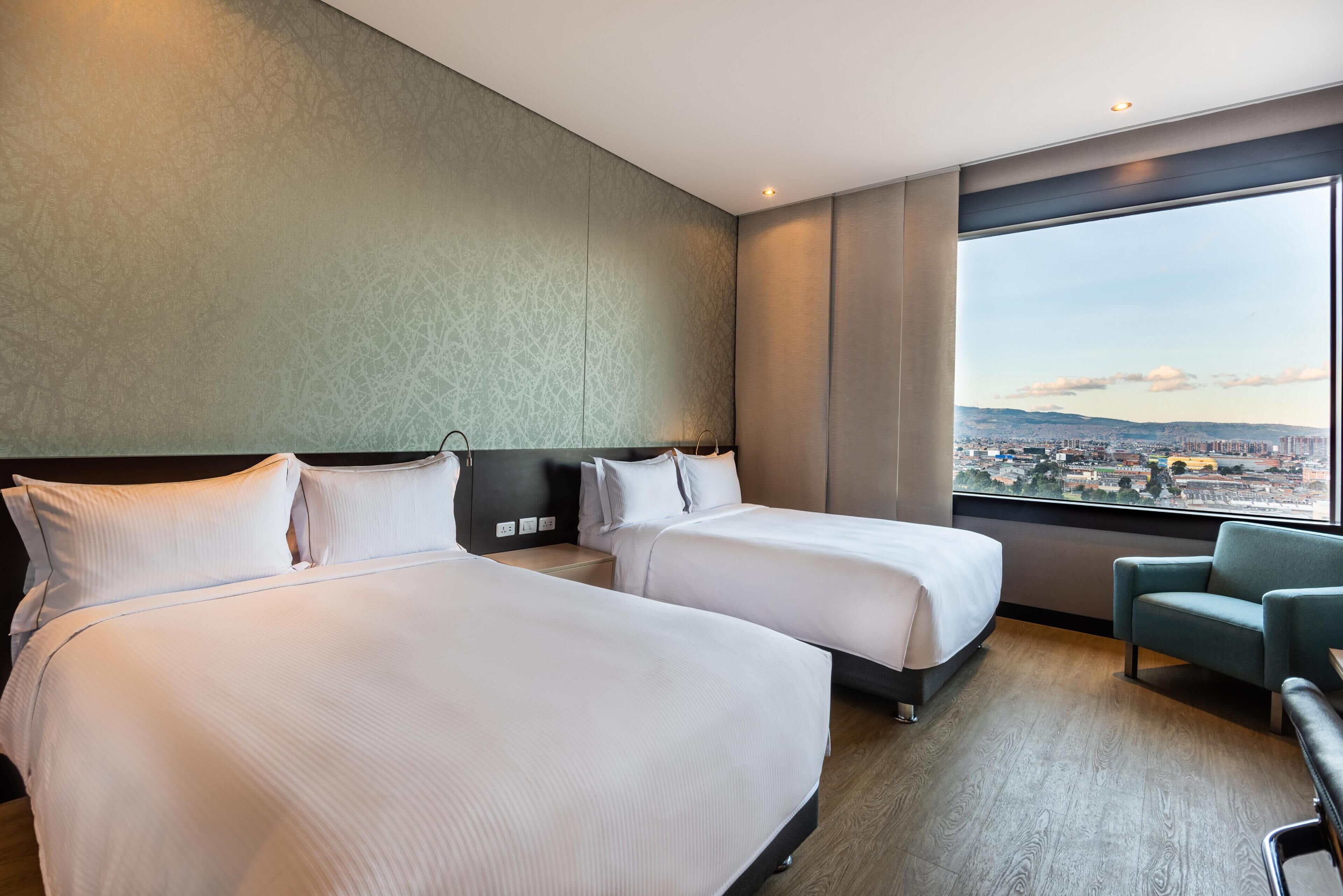 A hotel room with two beds, a view of a cityscape, and a green chair.