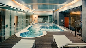 Un área de piscina interior moderna con paredes de vidrio, dos jacuzzis y sillones de descanso.