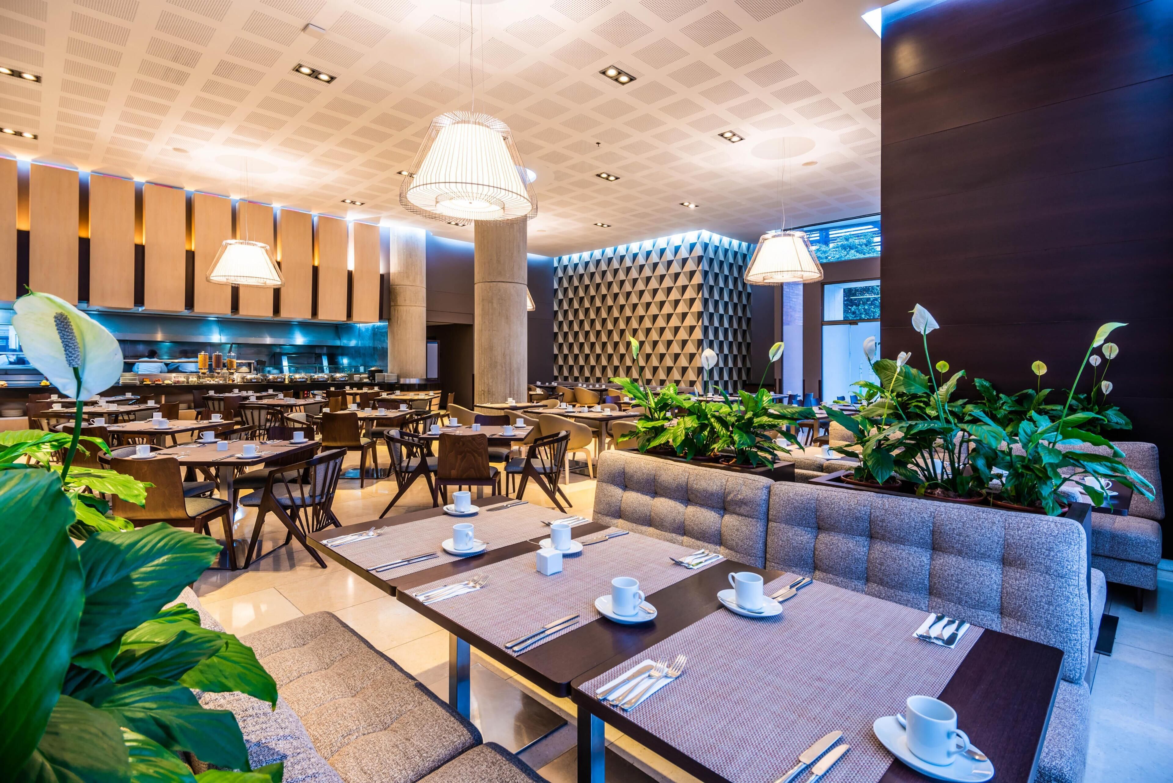 A modern dining area with tables set for service, a kitchen in the background, and a patterned wall.