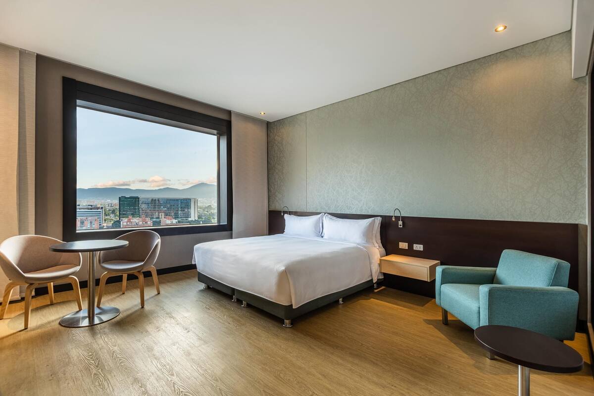 A hotel room with a large window, a bed, two chairs, a small table, and a nightstand.