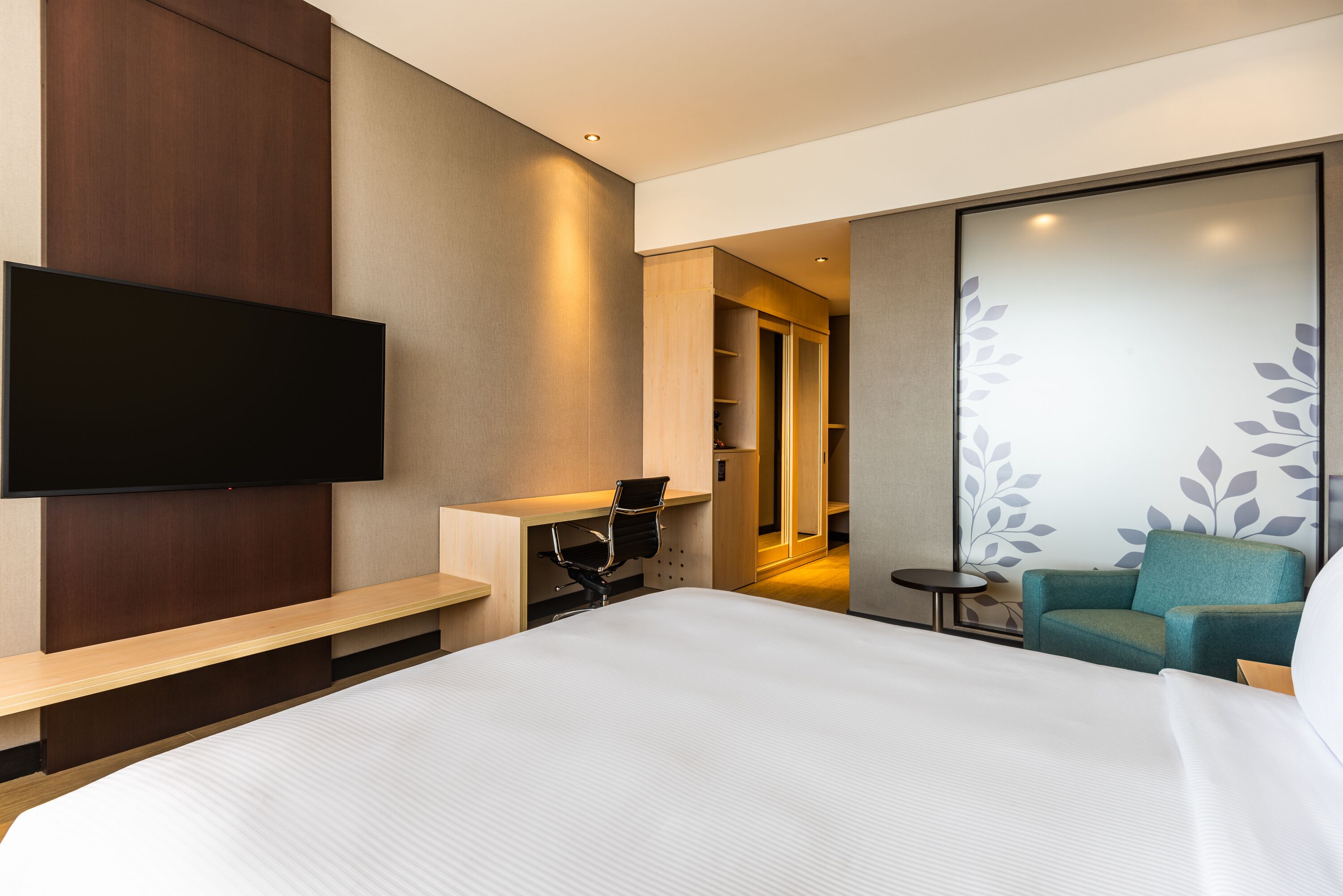 A modern hotel room with a large bed, a flat-screen TV mounted on the wall, a desk with a chair, a small table, and a sofa.