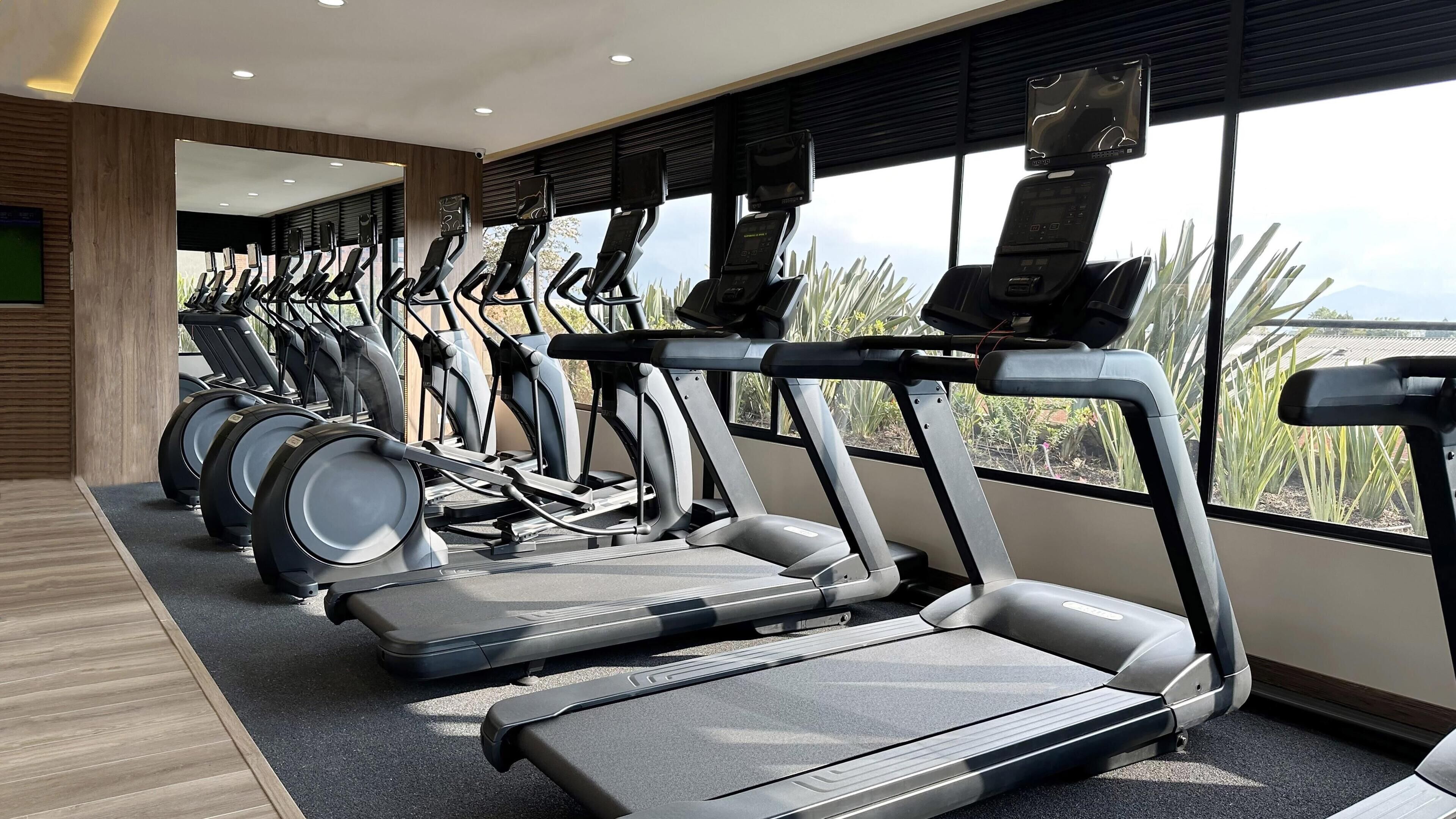 Un gimnasio con cintas de correr y bicicletas estáticas, amplias ventanas con vistas a zonas verdes y un diseño interior moderno.