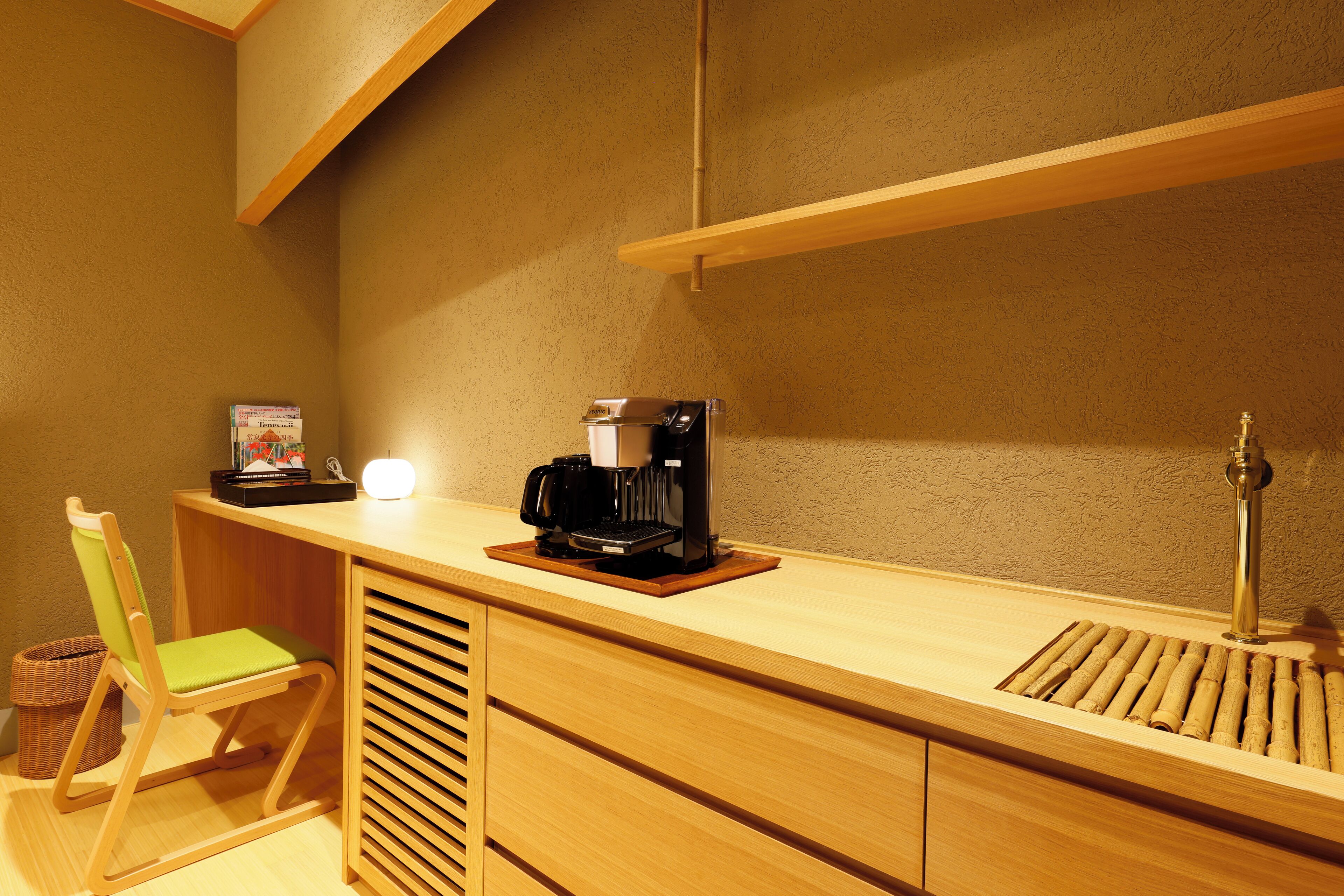 A compact room with a wooden desk, a coffee machine, a chair, and a small shelf.