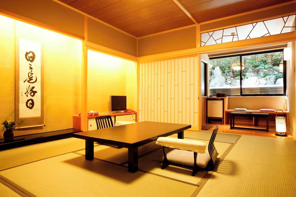 Ett traditionellt japanskt rum med tatamimattor på golvet, ett lågt bord, en bänk, en tv och ett fönster med utsikt över stenar och grönska.