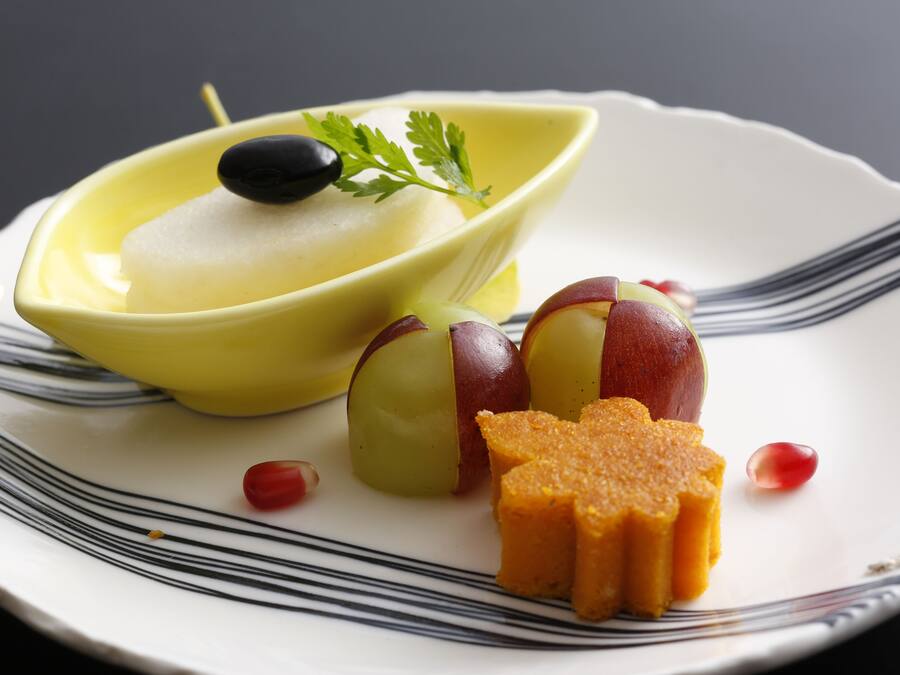 Un plato con un tazón amarillo que contiene una sustancia blanca, dos manzanas cortadas por la mitad con pulpa roja, y una porción de pastel de naranja.