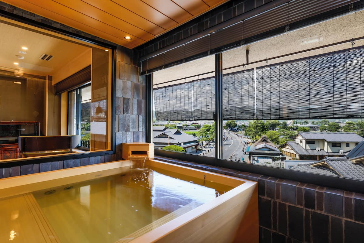 Ett traditionellt japanskt onsen-badhus med ett träbadkar, stora fönster och utsikt över traditionella byggnader.