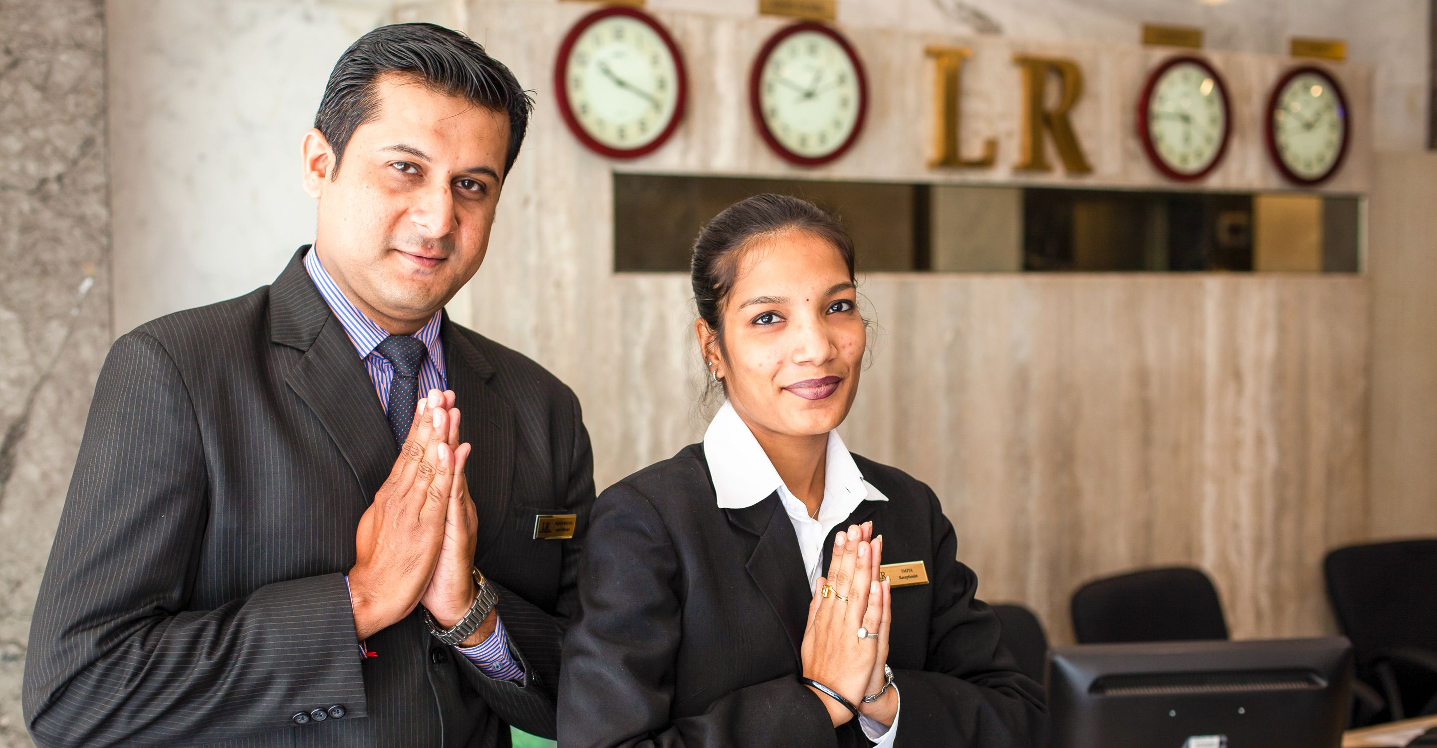 Two individuals in business attire standing in a lobby with clocks and a sign that reads "LRO".