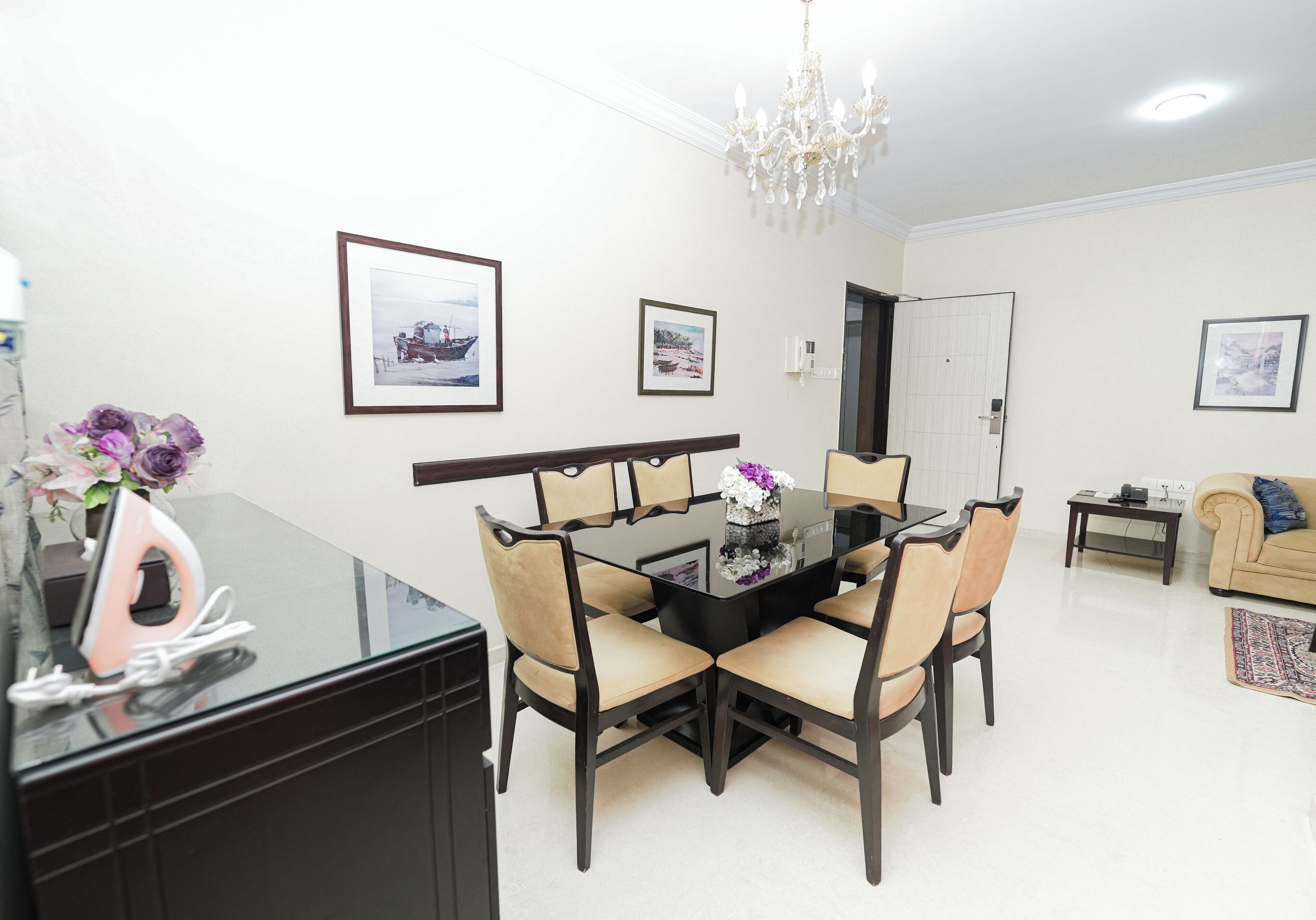 A dining area with a glass table, four chairs, a chandelier, and framed pictures on the walls.