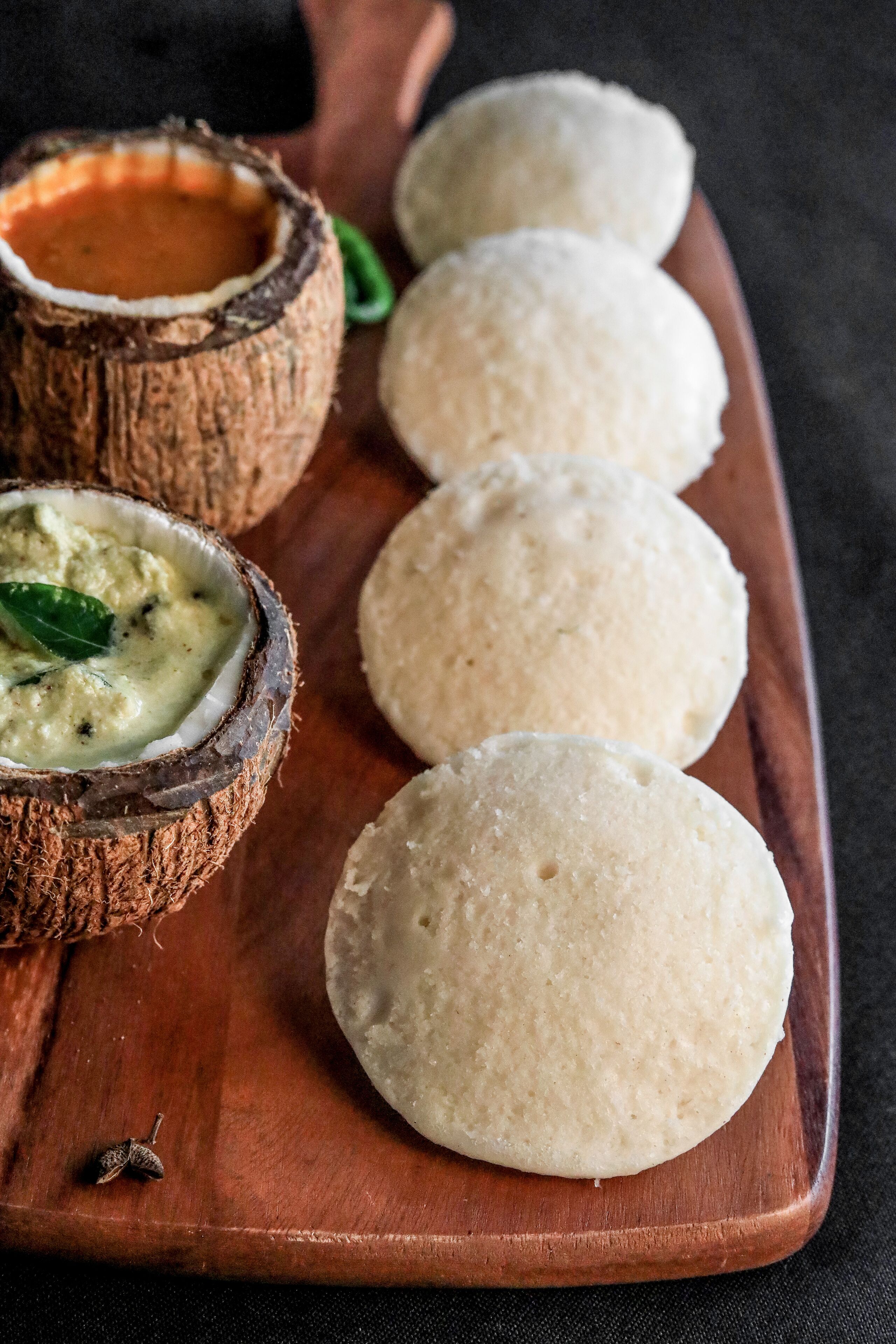 A wooden serving board with three round, white, fluffy food items and a coconut shell containing a creamy sauce with herbs.
