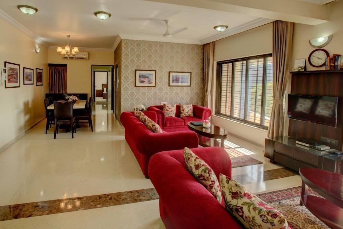 A spacious living area with a red sofa set, a dining table with chairs, a TV, and a large window with curtains.