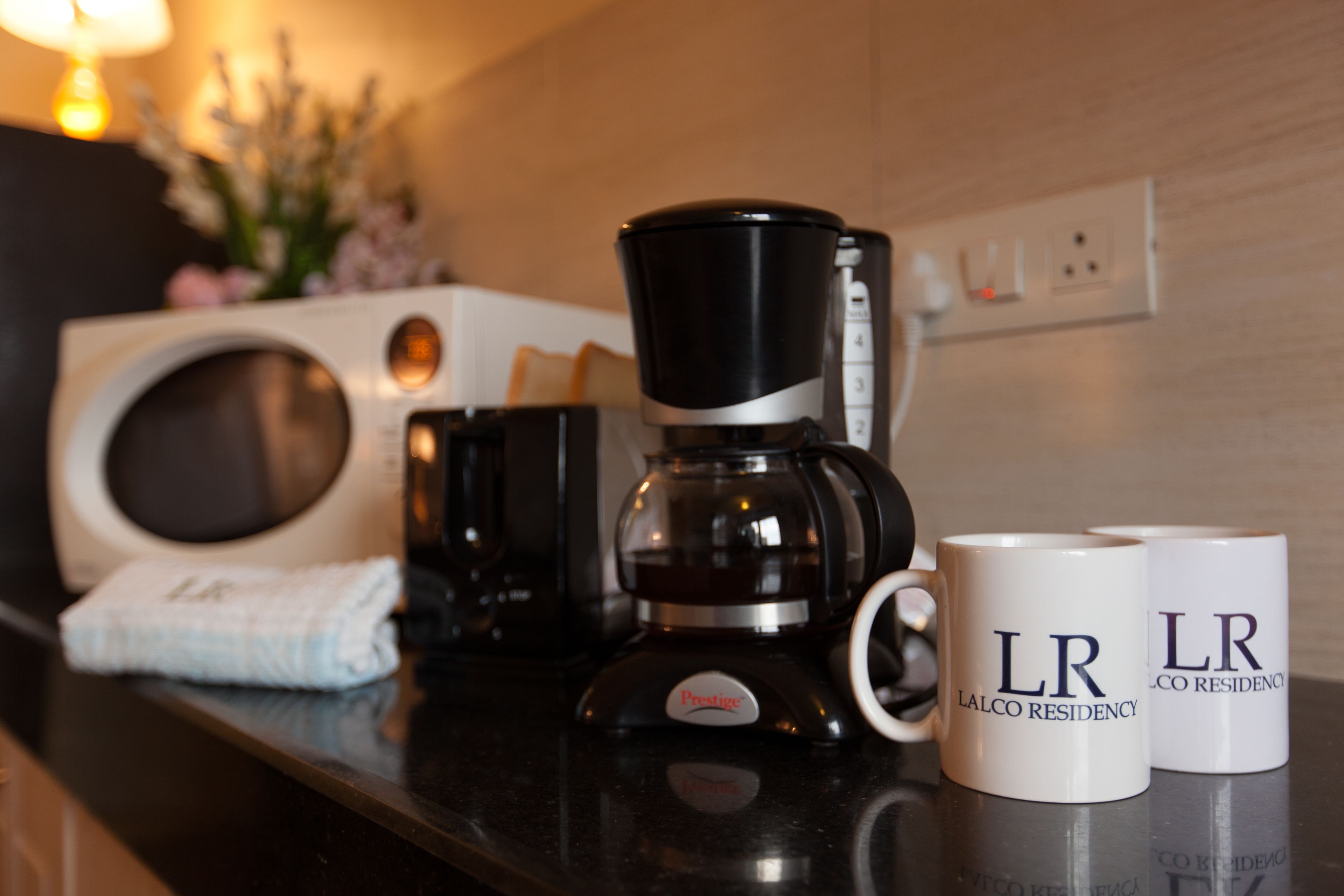 A coffee maker, two mugs with "LR LALCO RESIDENCY" text, a towel, and a microwave on a kitchen counter.