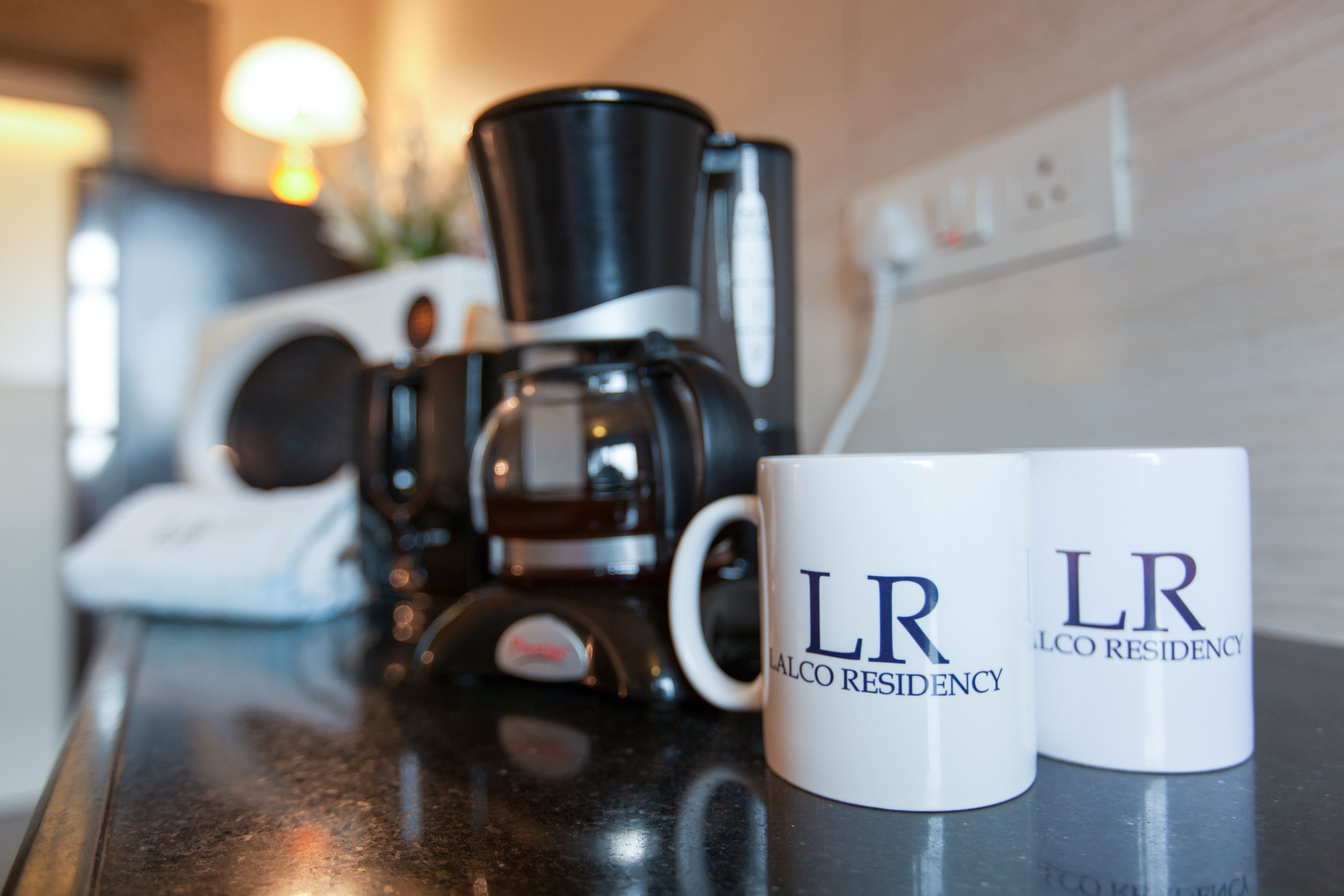 A coffee maker and two mugs with "LR" and "ALCO RESIDENCY" on a countertop.